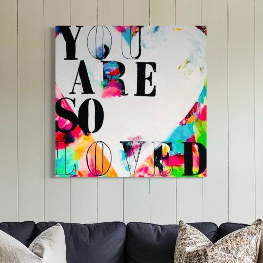 Colorful wall art with 'YOU ARE SO LOVED' text in a living room.