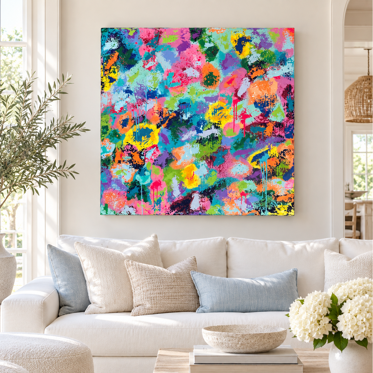 A vibrant, multicolored abstract canvas painting is displayed on a light beige wall above a modern white sofa with neutral throw pillows in a bright, contemporary living room.