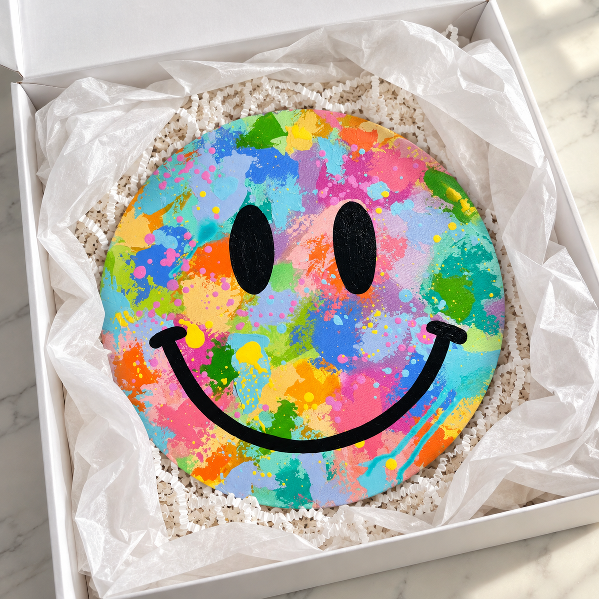 A round decorative item with a colorful paint-splatter smiley face design, presented inside a white gift box with tissue paper.