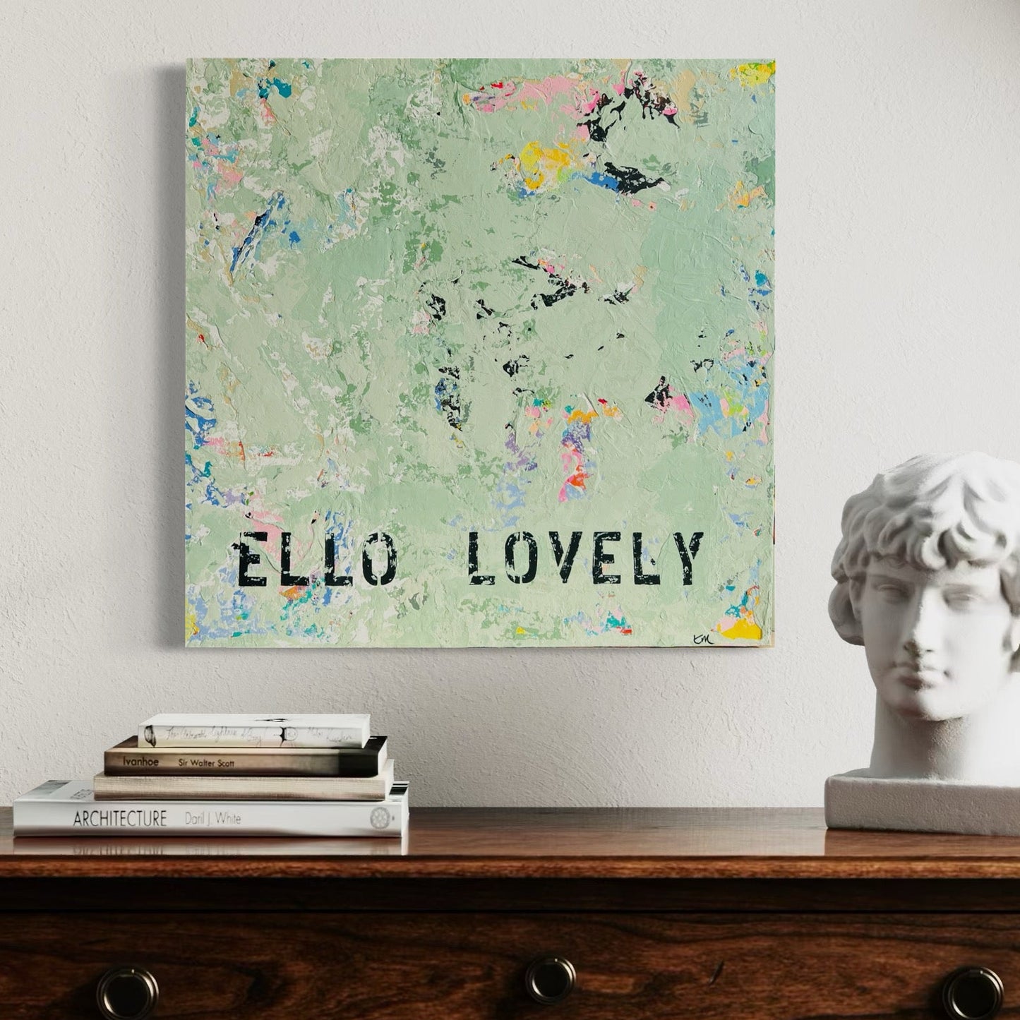 Decorative wall art with 'ELLO LOVELY' text on a floral background, displayed on a wooden surface with books and a statue.
