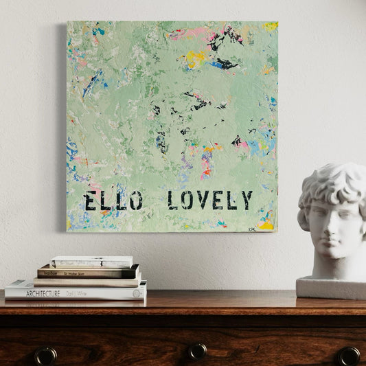 Decorative wall art with 'ELLO LOVELY' text on a floral background, displayed on a wooden surface with books and a statue.