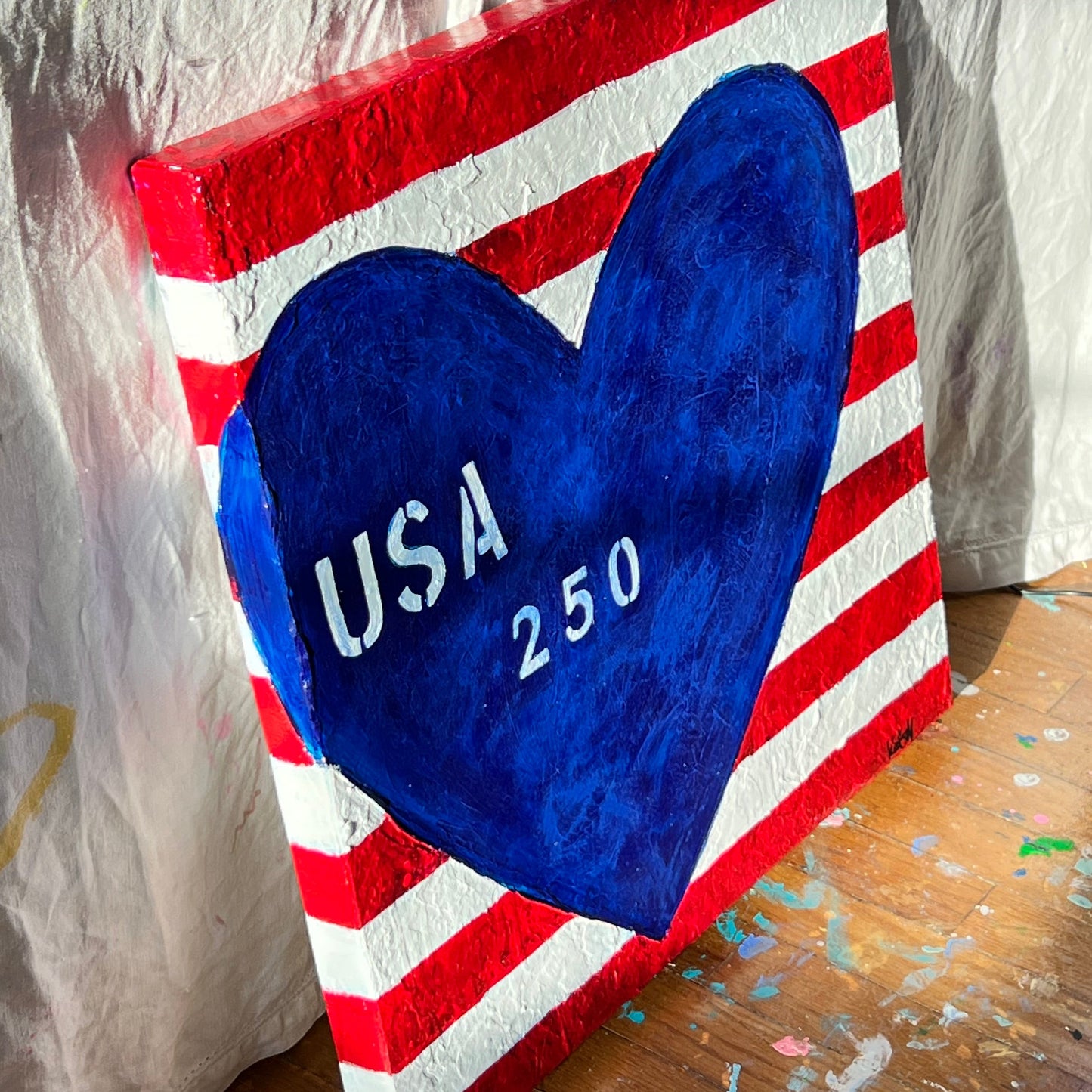 Canvas with a blue heart and 'USA 250' text on a red and white striped background, leaning against a wooden wall.